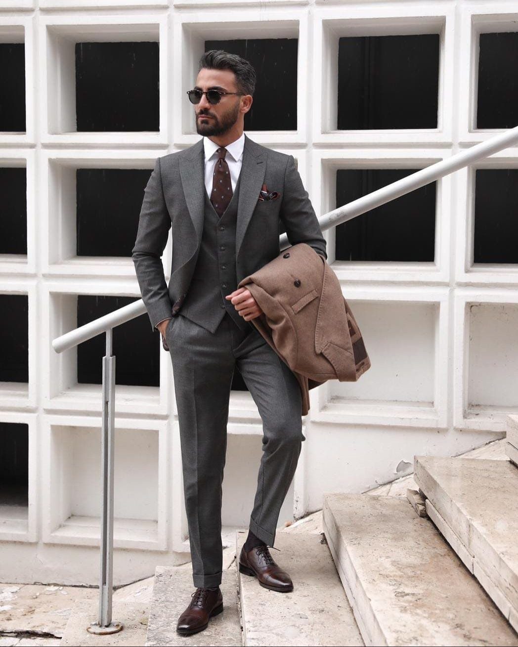 Man in a grey three-piece suit and sunglasses standing on outdoor stairs.