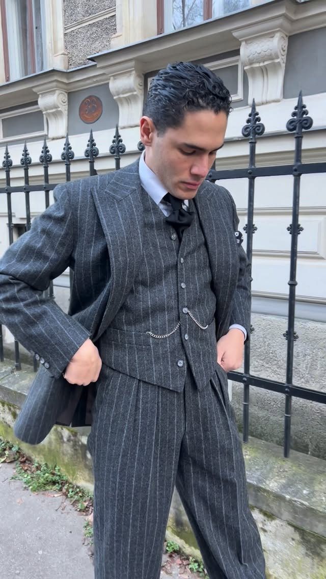 A man wearing a dark grey pinstripe three-piece suit with a silver pocket watch chain.