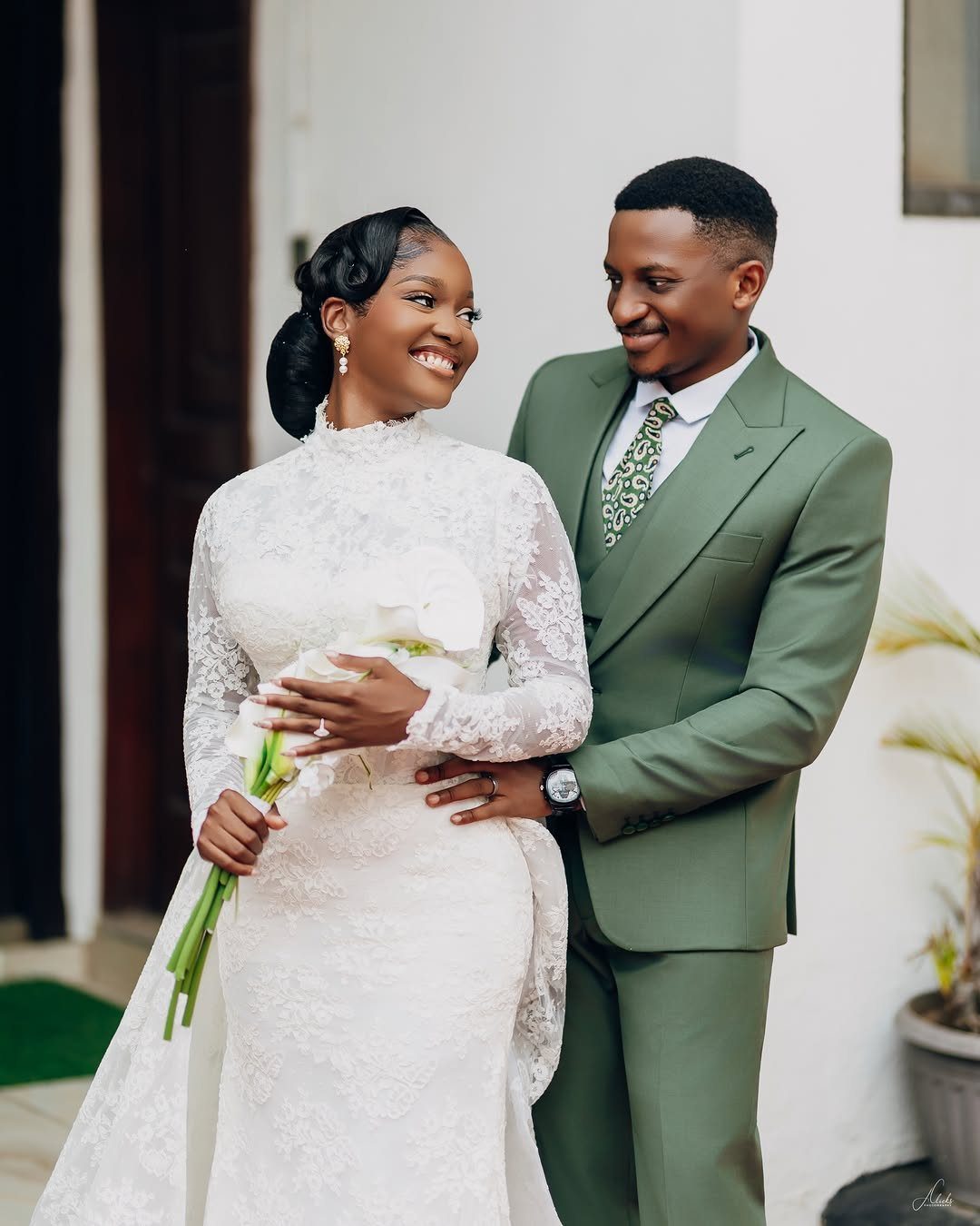 A man in a sage green three-piece suit smiling at a woman in a white lace dress.