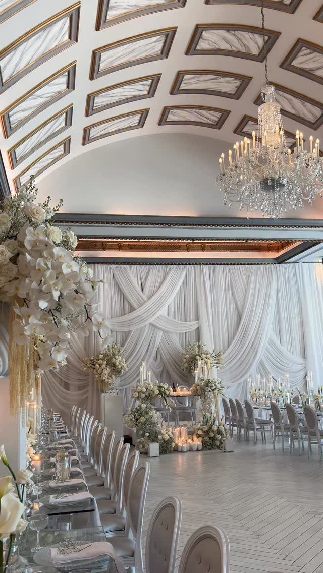 White draped fabric and floral arrangements decorate an elegant ballroom with a crystal chandelier.