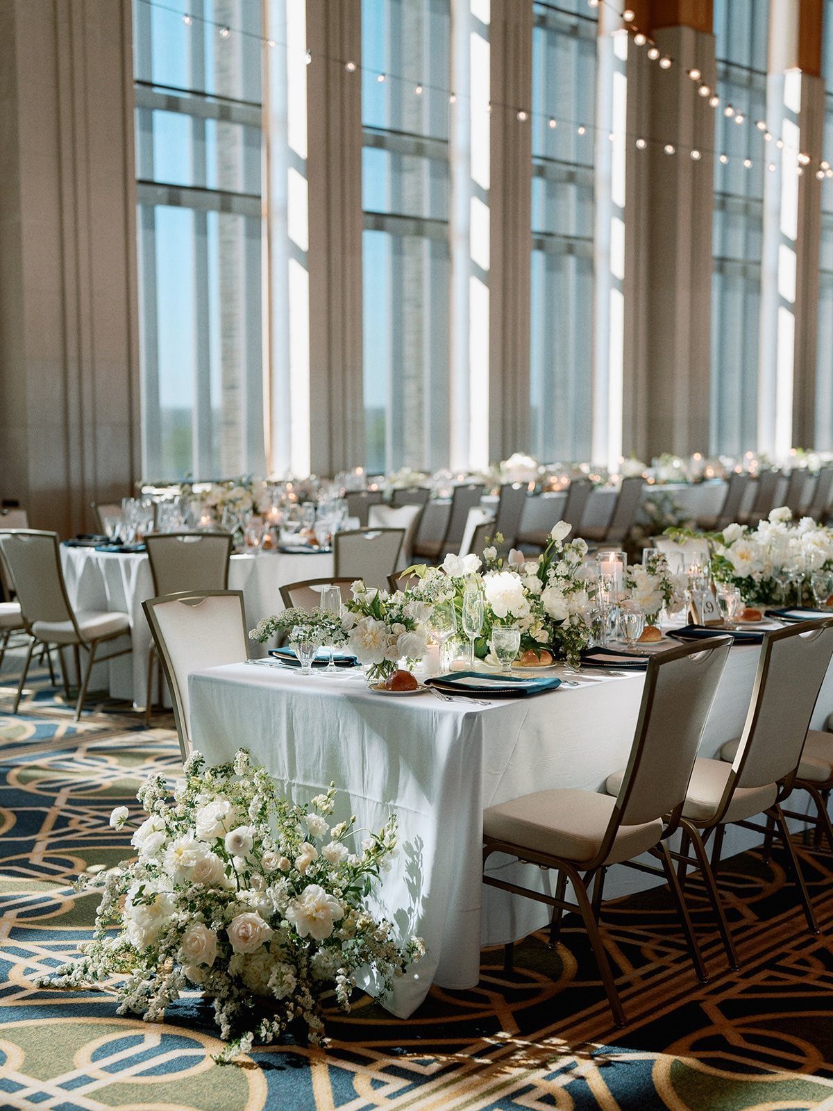 Set banquet tables with white linens and floral centerpieces in a bright, large ballroom venue.