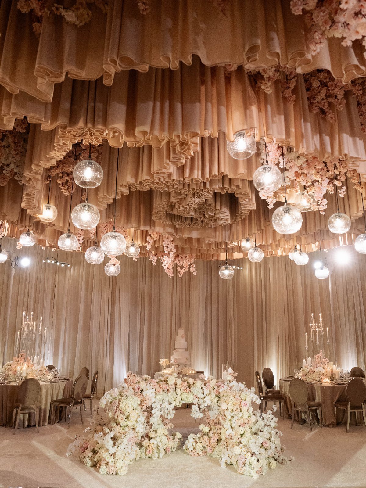 Elegant ballroom wedding reception with draped peach ceilings, hanging globe lights, and lush floral arrangements.
