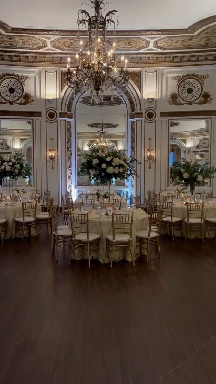 An elegant ballroom decorated with round tables, gold chairs, and large floral centerpieces under a chandelier.