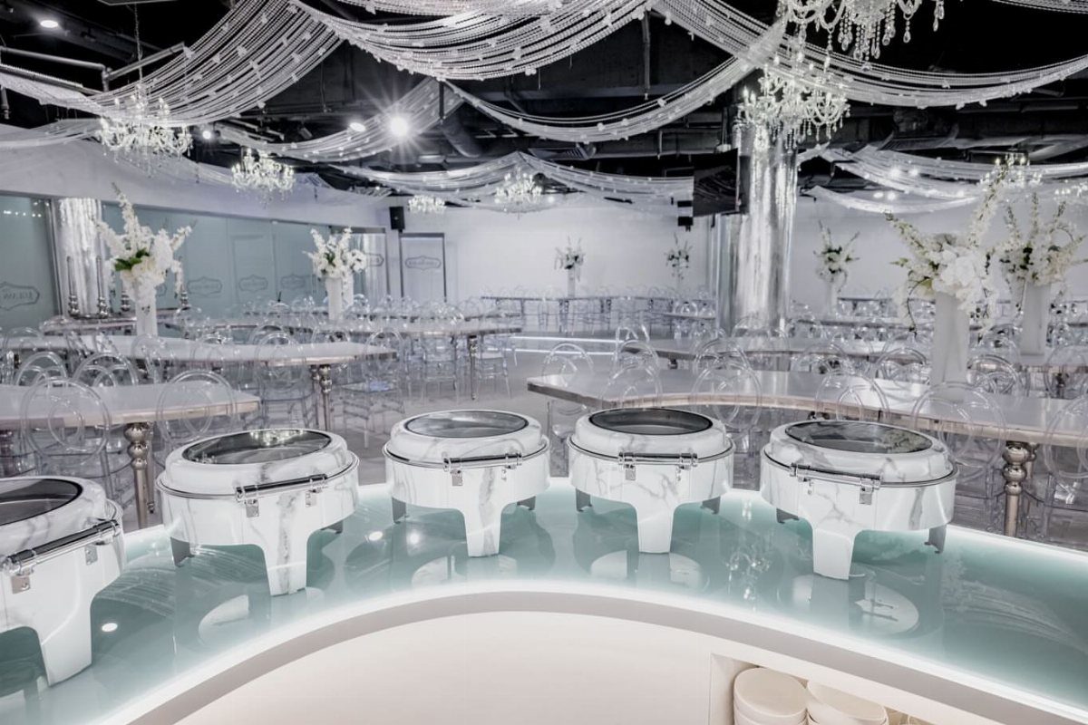 White marble chafing dishes arranged on a glowing buffet counter in a decorated ballroom.