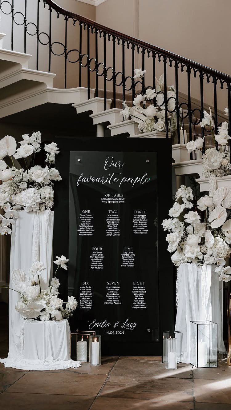 A black wedding seating chart with white calligraphy stands between white floral arrangements near a staircase.