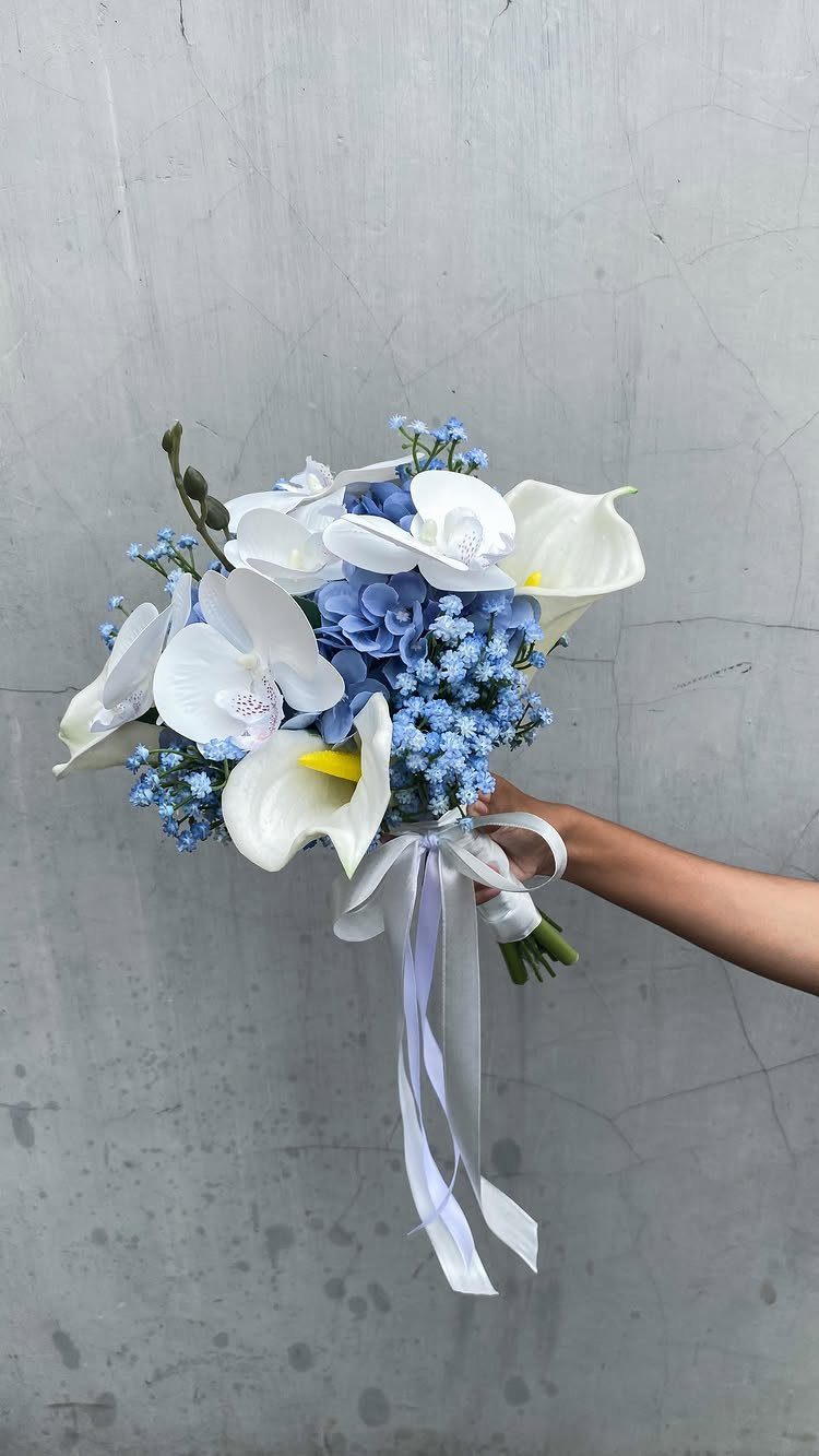 A hand holding a wedding bouquet with white lilies, white orchids, and blue hydrangea flowers.