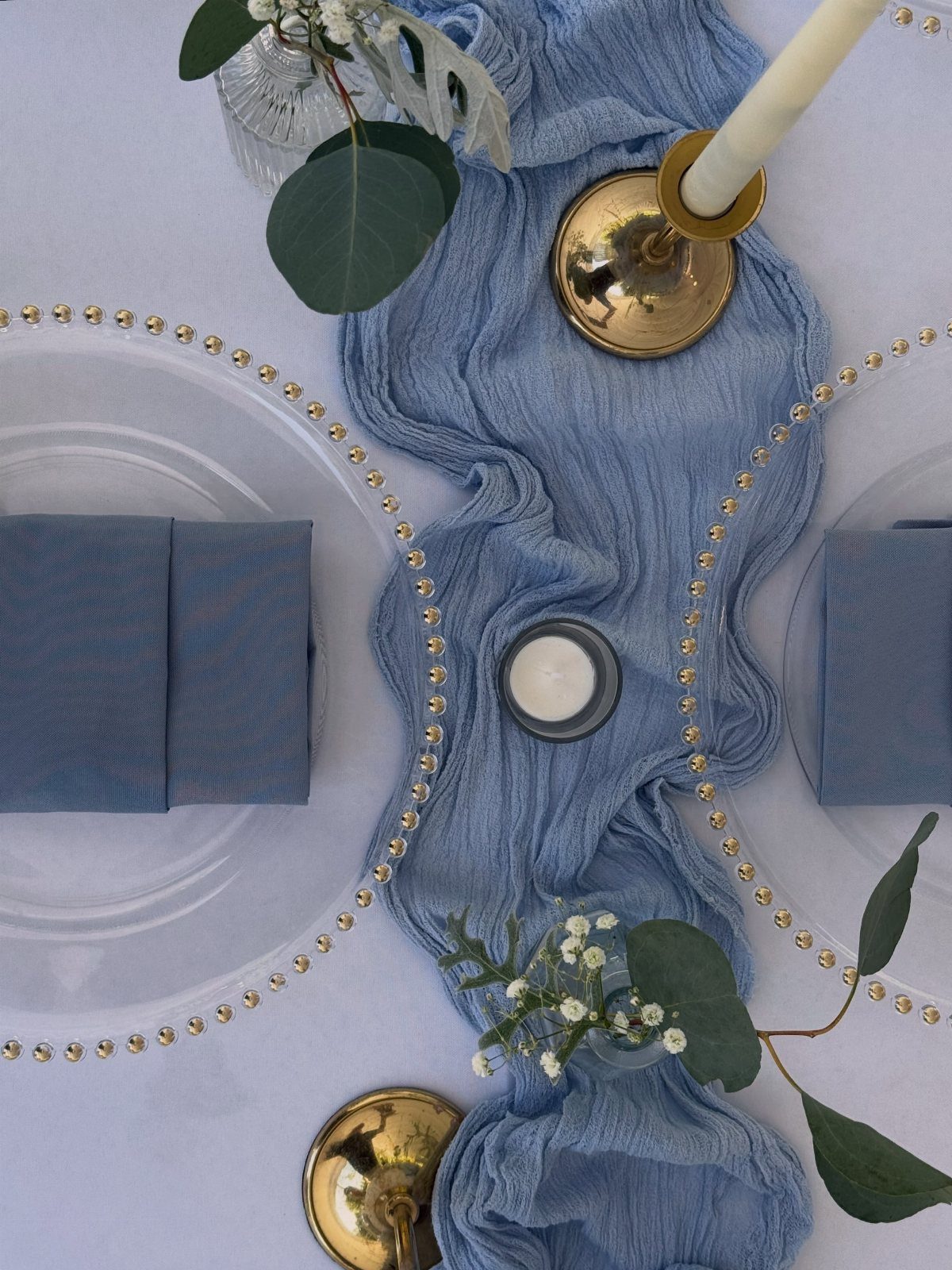 Dusty blue gauzy table runner on a white tablecloth with plates, napkins, and brass candle holders.
