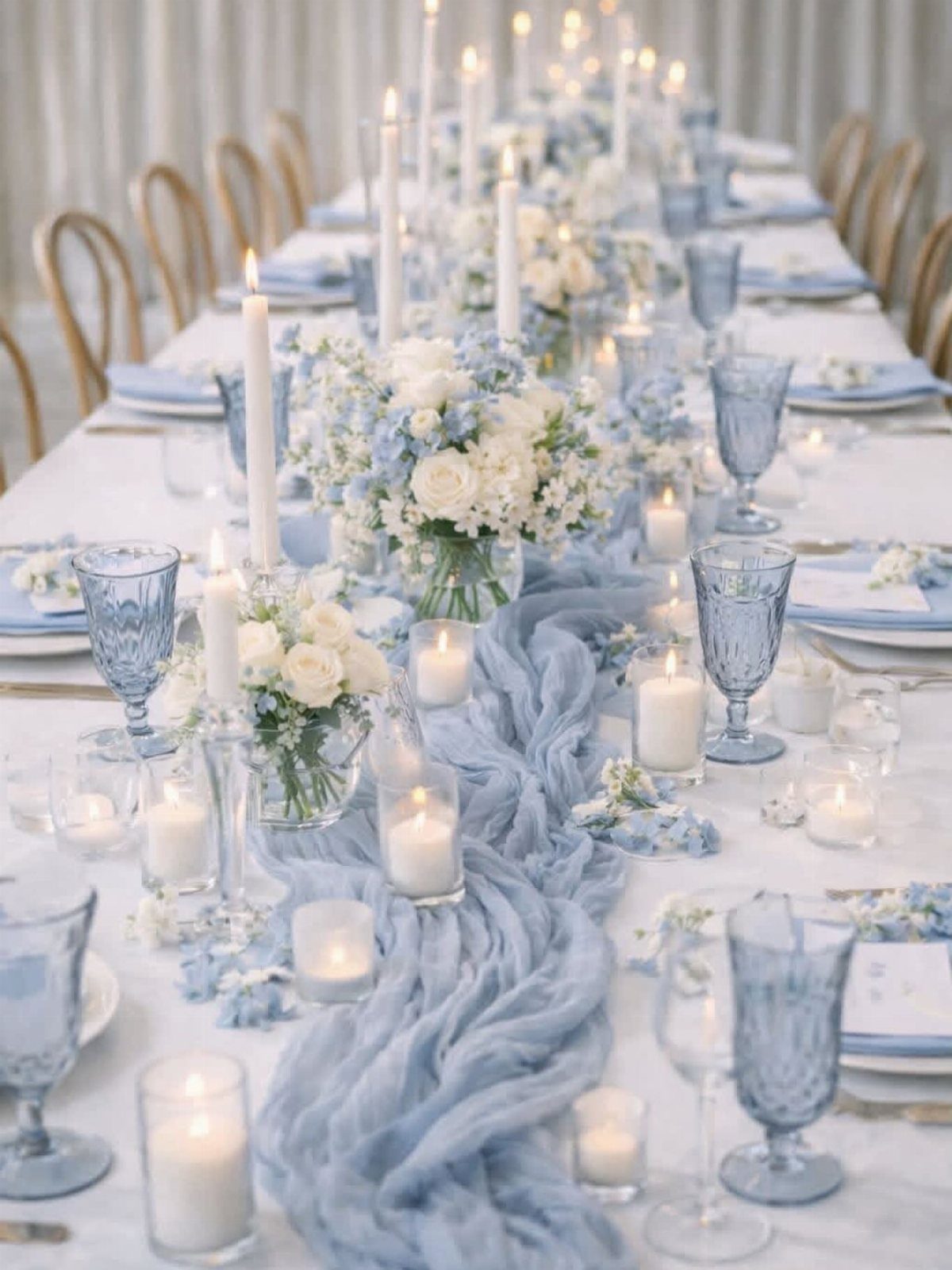 A long wedding reception table decorated with dusty blue gauze runners, white flowers, and many glowing candles.