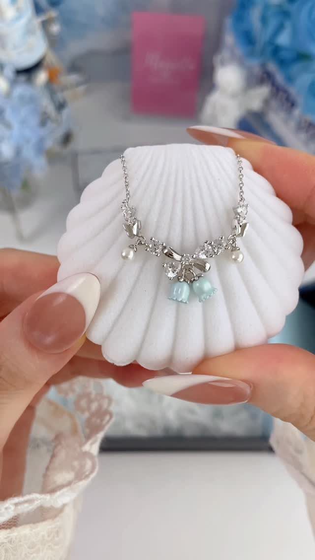 A silver crystal necklace with light blue beads resting on a white seashell holder held by hands.