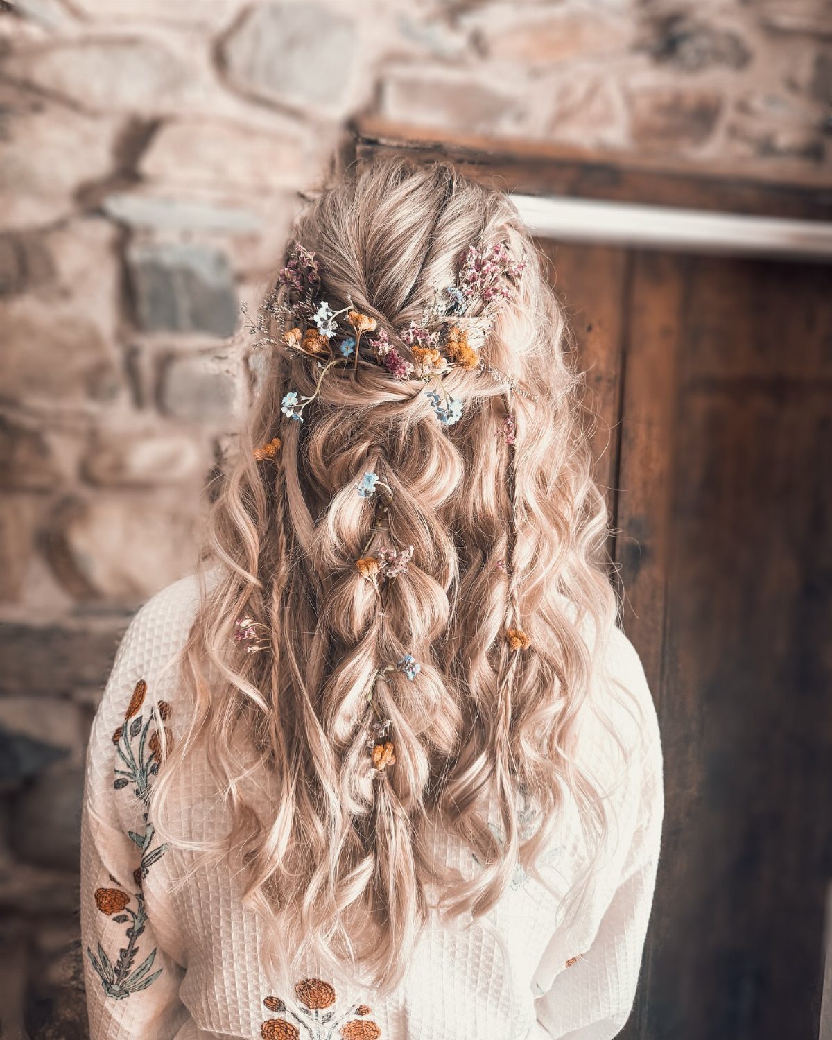 Long wavy blonde hair styled with a braid and small colorful dried wildflowers.