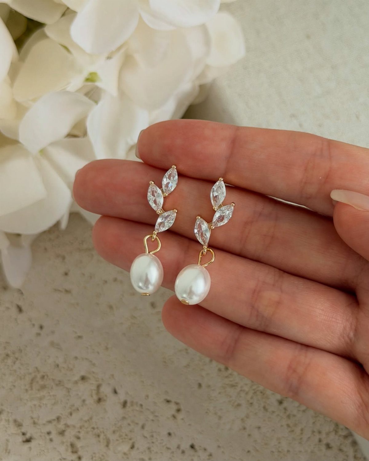 Bridesmaid Jewelry Gold: 10 Ideas for Every Wedding Style 5 A hand holding gold drop earrings with marquise crystals and white pearls against white flowers.