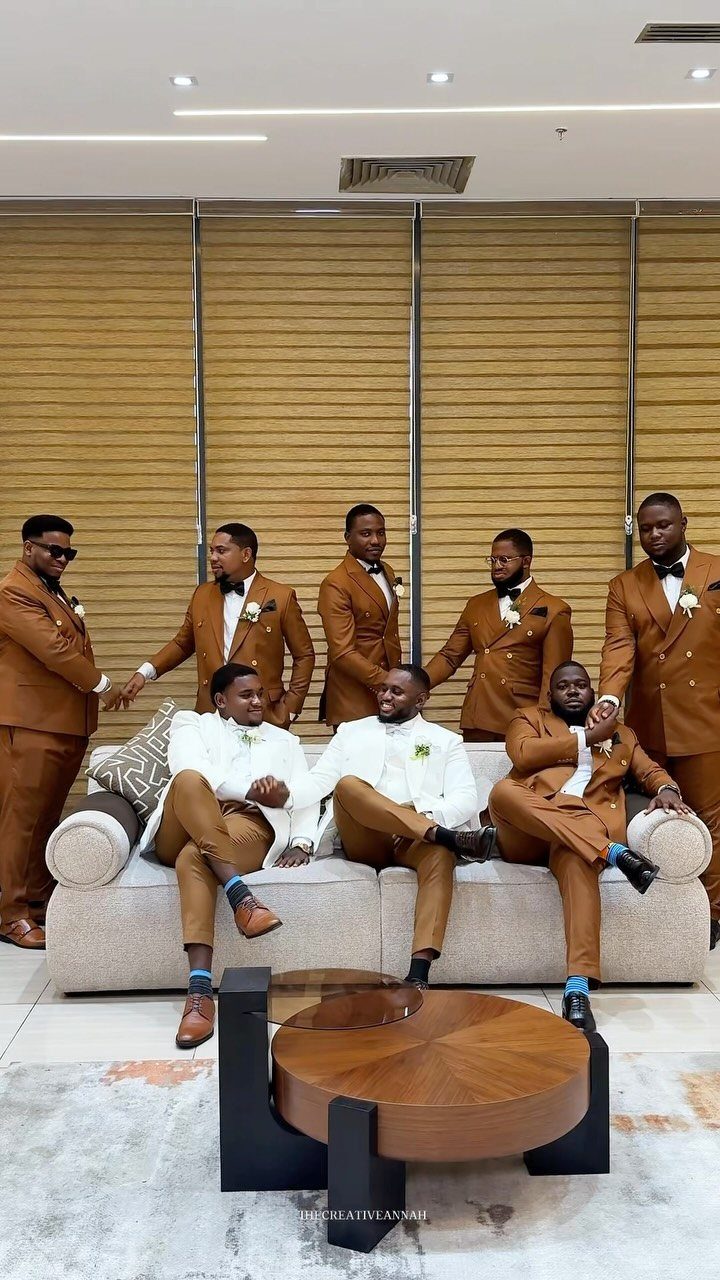 Seven men posing in matching brown tailored suits and white shirts in a bright room.