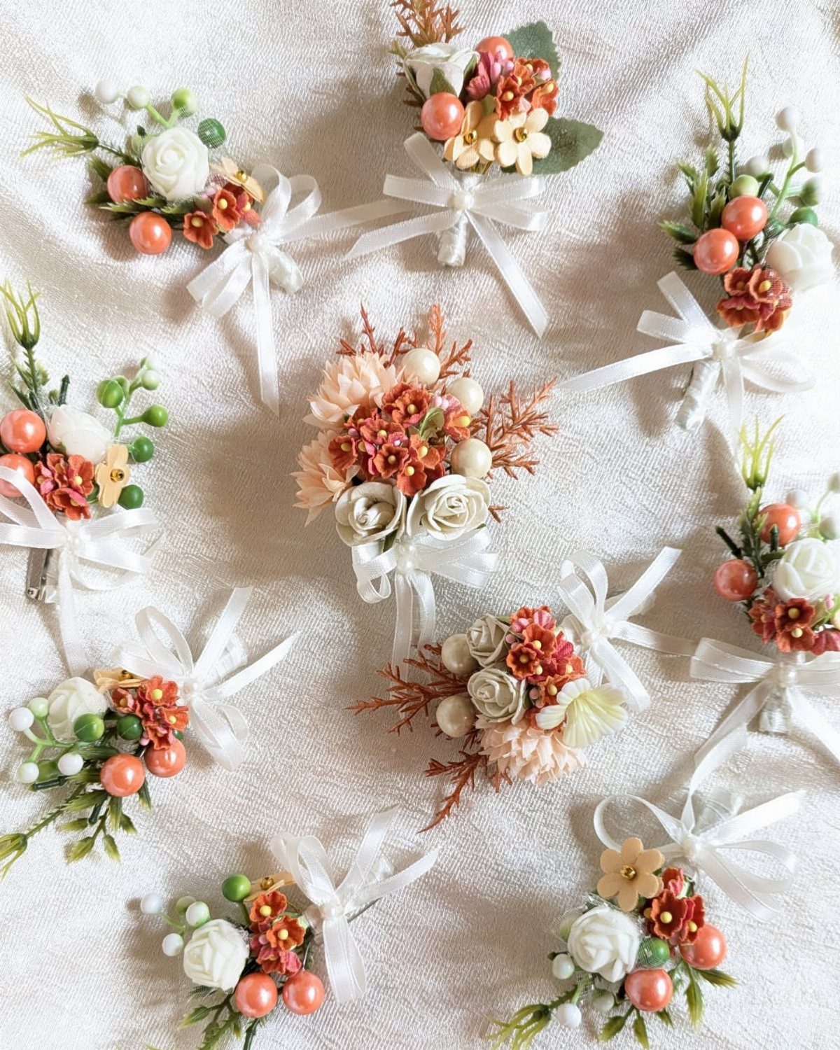 Multiple small floral boutonnieres with peach and white flowers tied with white ribbons on white fabric.