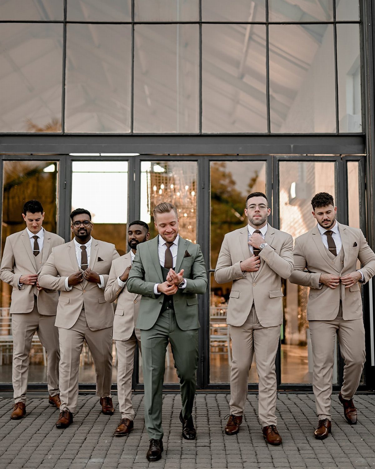 Six men posing in light beige suits and dark brown ties in front of a glass building.