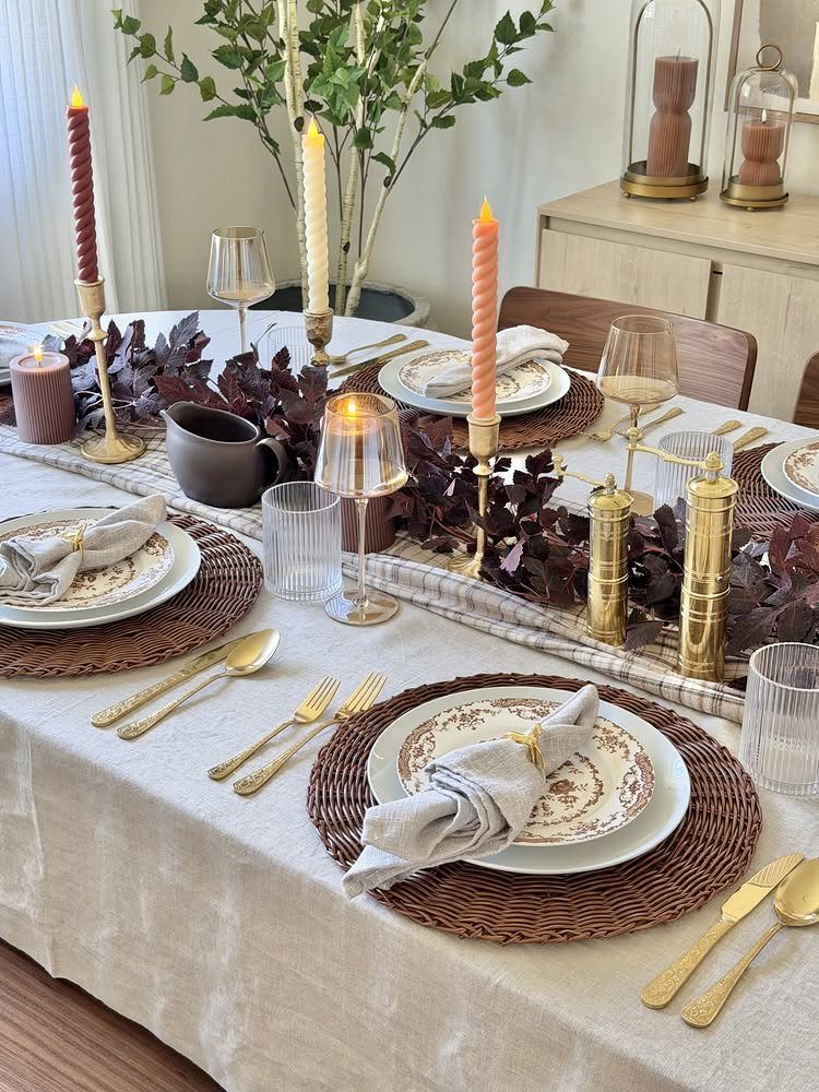 A formal table setting featuring patterned plates, brass cutlery, and tall taper candles on linen.