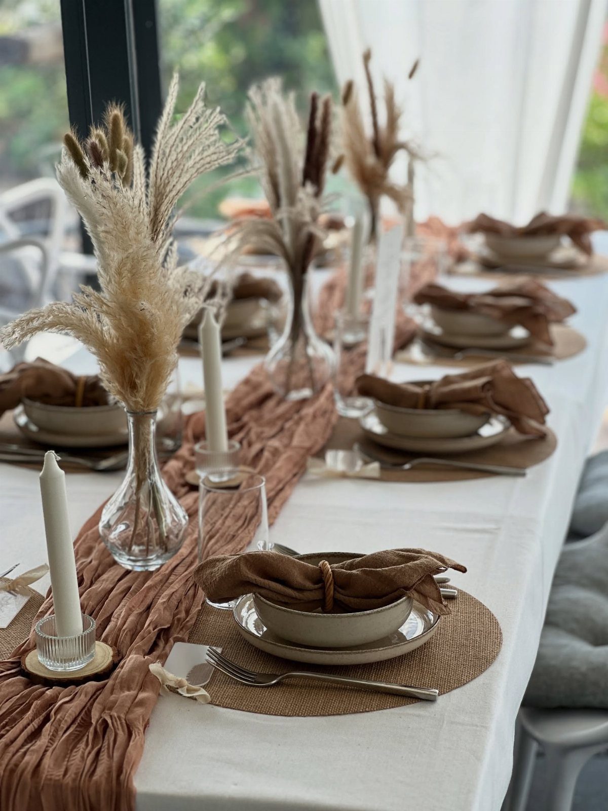 A dining table set with beige plates, brown napkins, and dried pampas grass in glass vases.