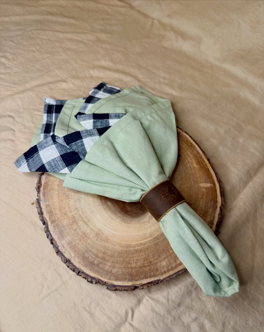 A light green and checkered napkin held by a brown leather ring on a wood slice.