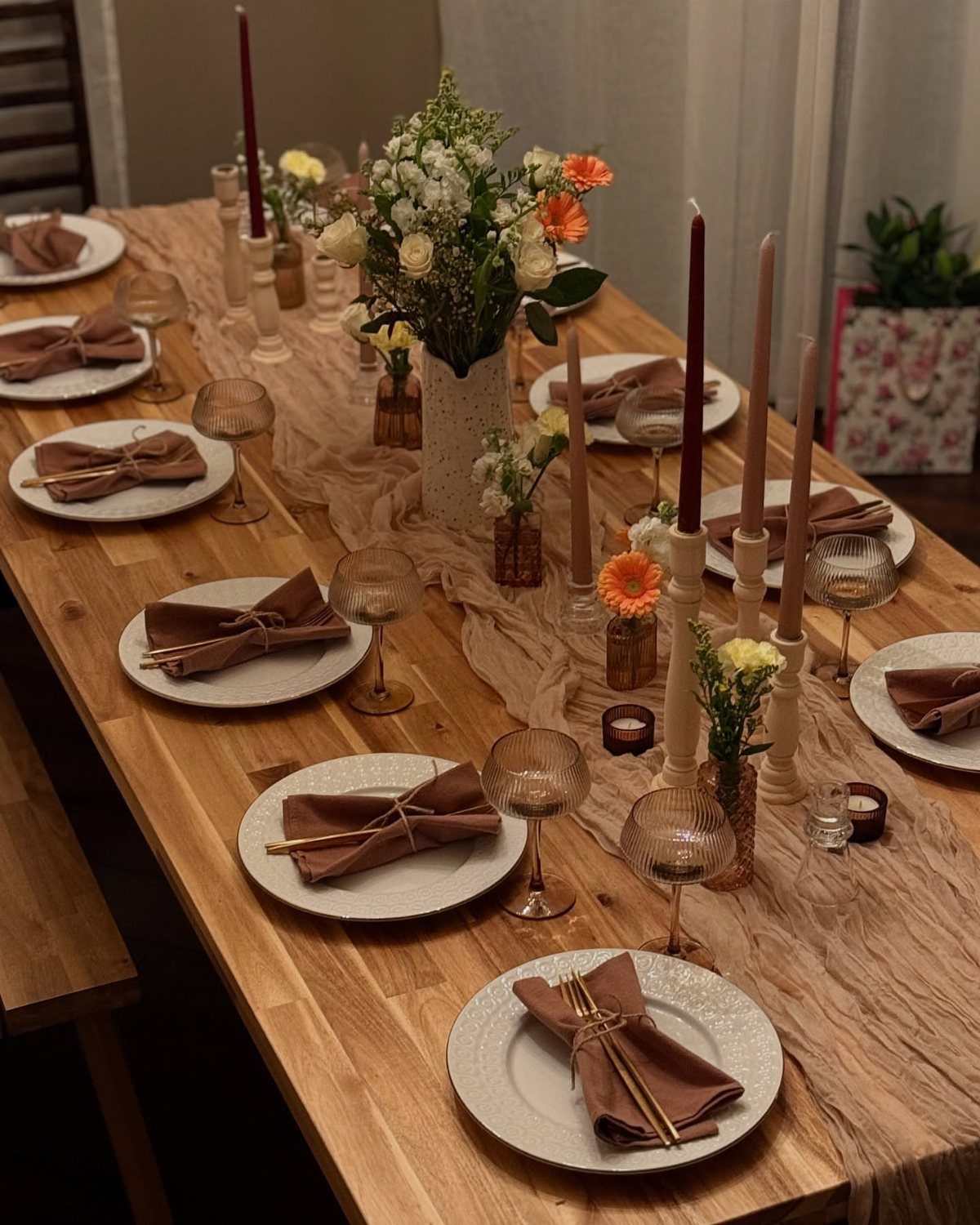 A long wooden dining table decorated with white plates, brown napkins, amber glassware, and floral arrangements.