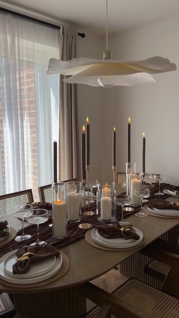 A long wooden dining table set with white plates, brown napkins, and various lit candles.