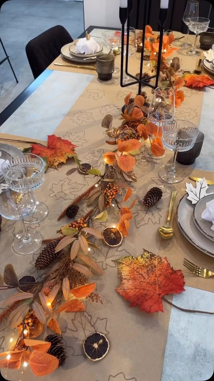 An autumn themed table setting with leaves, pinecones, and fairy lights on a brown runner.