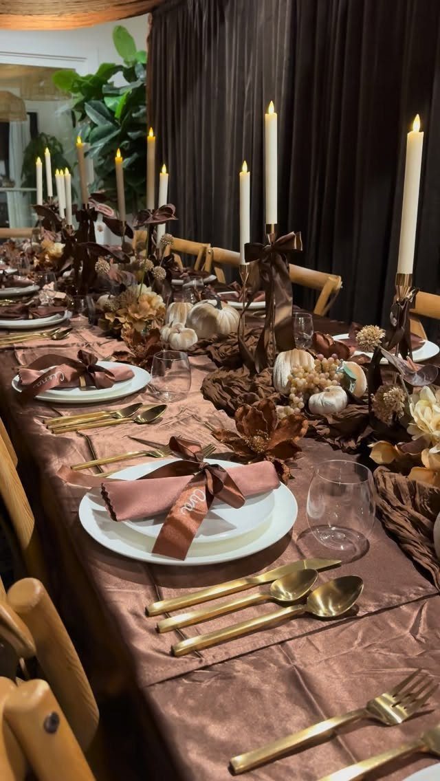 A long dining table decorated with brown linens, white plates, gold cutlery, and taper candles.