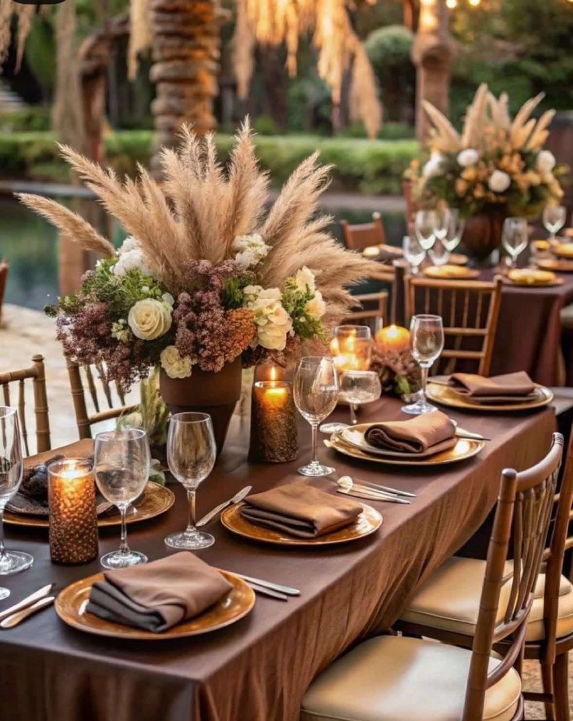 A formal dining table set with brown linens, gold chargers, and a large pampas grass centerpiece.
