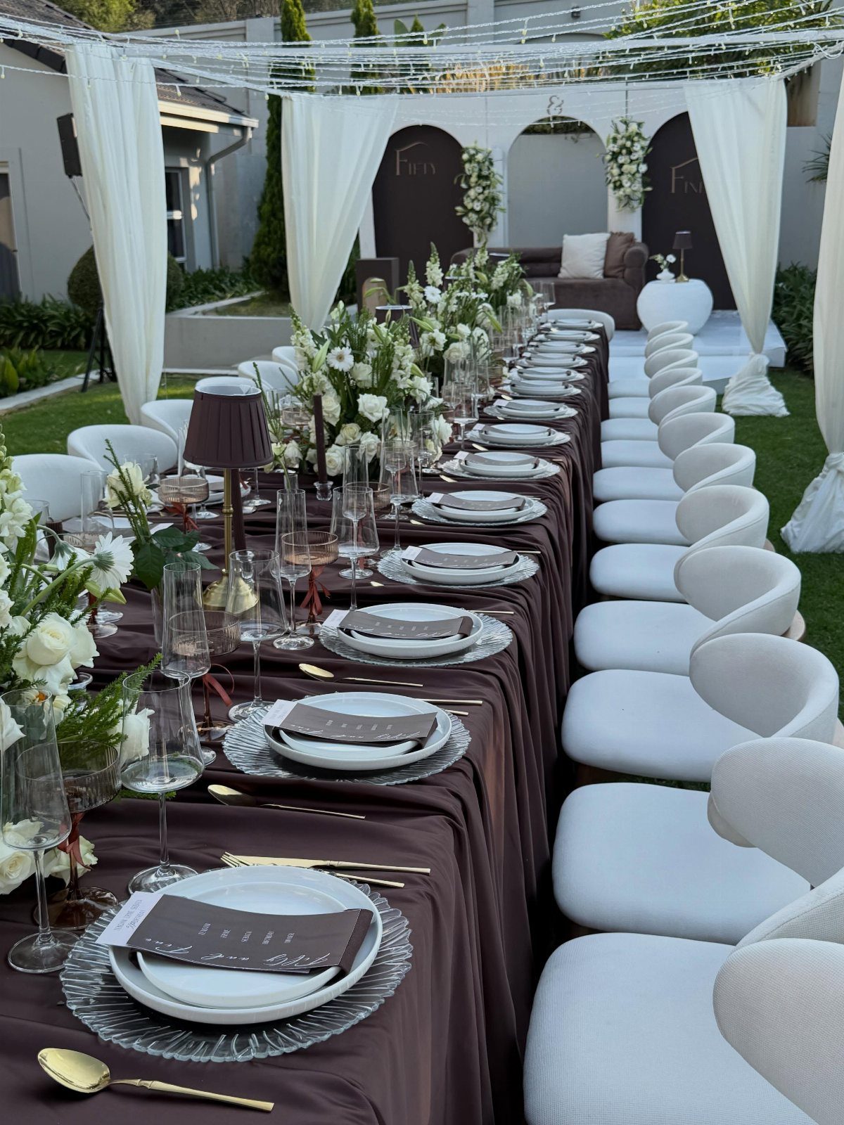 Long dining table with brown cloth, white plates, and white floral arrangements outdoors.