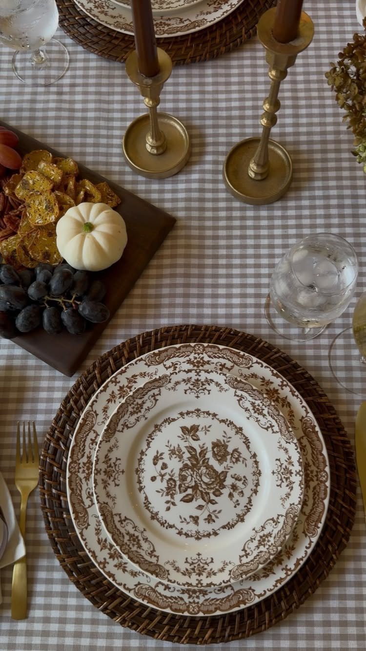 A table setting with gingham tablecloth, patterned plates, woven chargers, and brass candle holders.