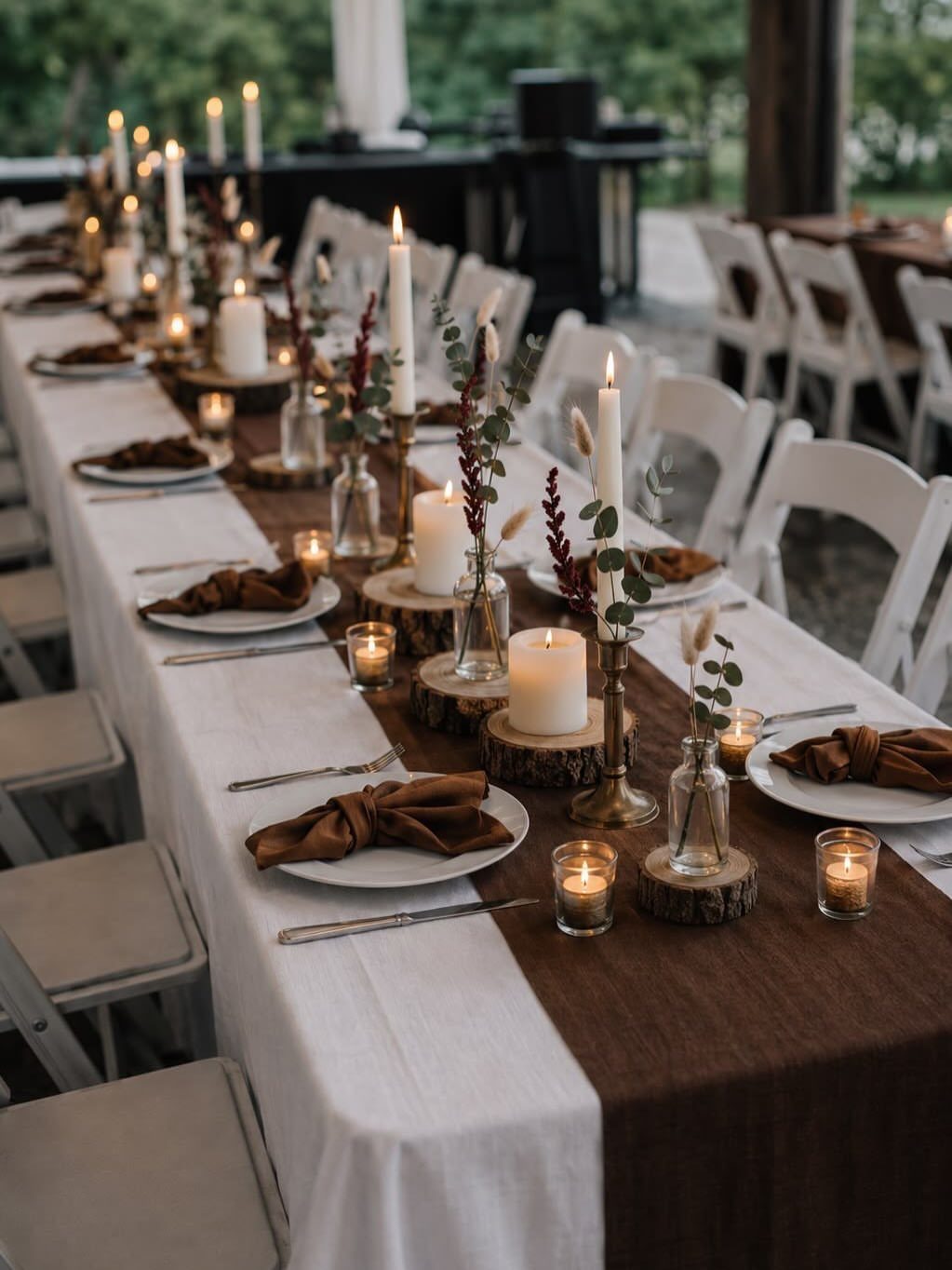 A long dining table set with white plates, brown napkins, and layered linens with candles.