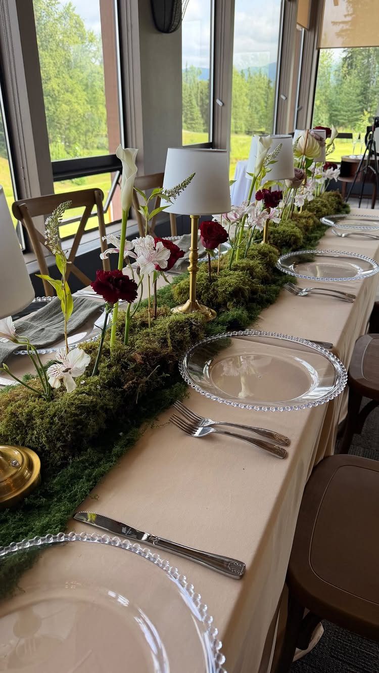 A long dining table featuring a lush green moss centerpiece with white and red flowers.