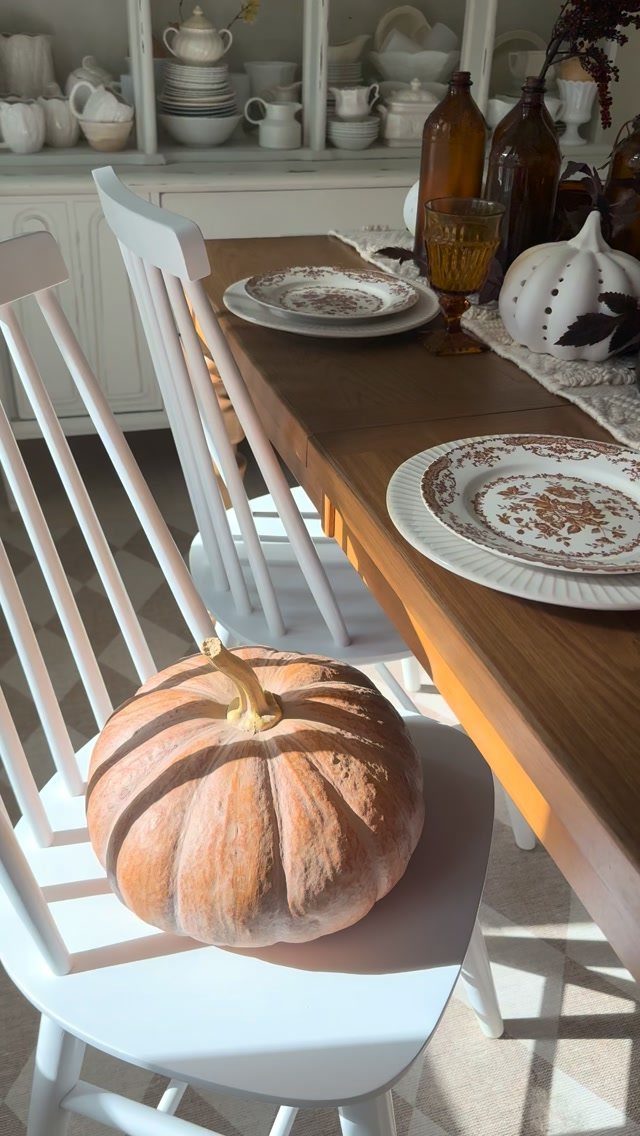 A wooden dining table set with patterned plates, amber glassware, and a large pumpkin on a white chair.