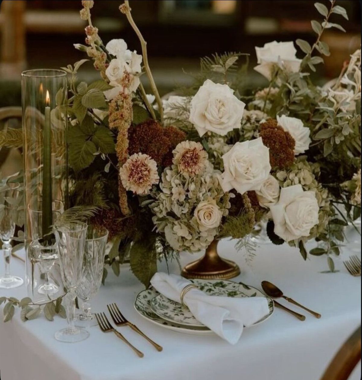 White roses and dried flowers in a gold vase on a white table setting.