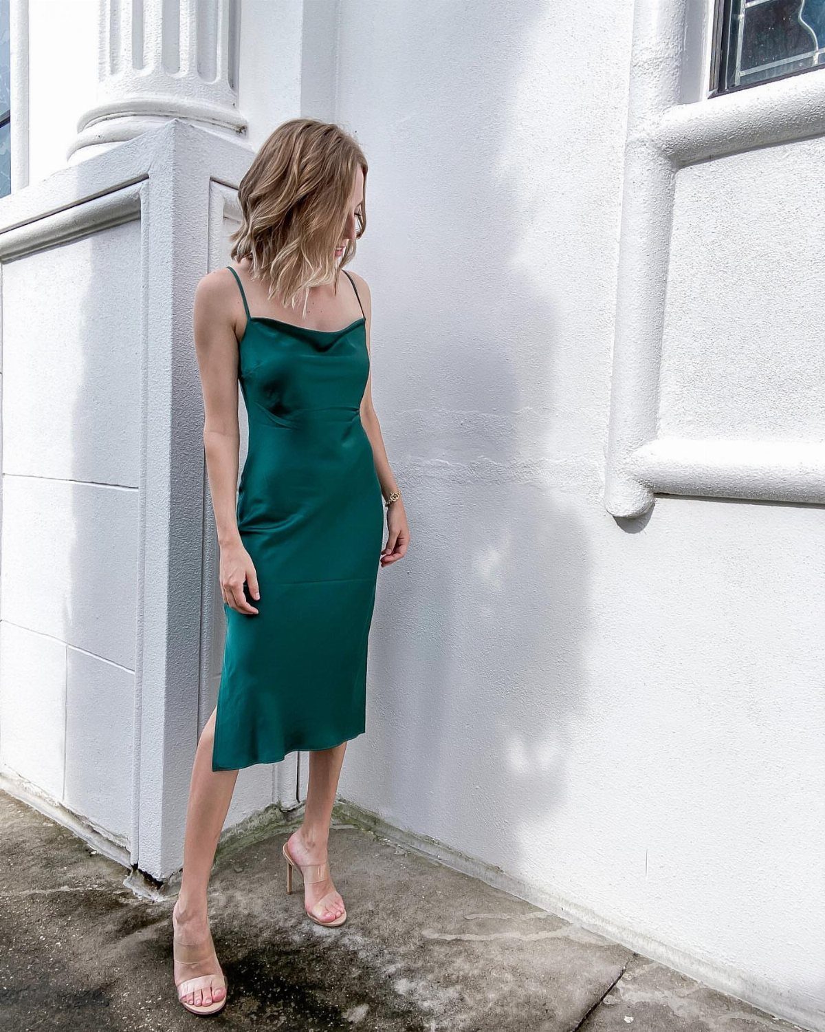 A woman wearing a dark green satin slip dress and clear heels standing against a white wall.