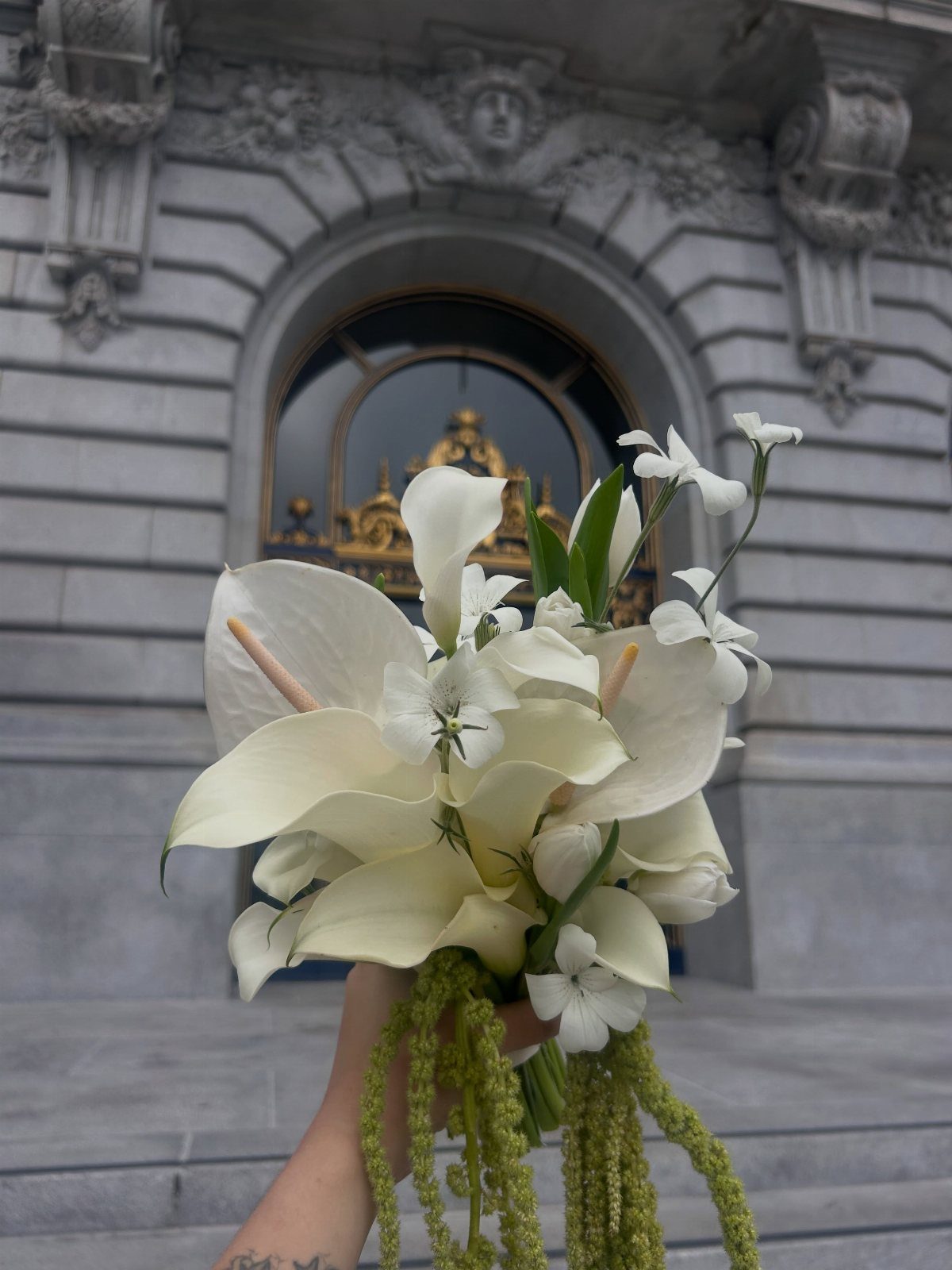 A hand holding a white anthurium and jasmine bouquet with trailing green amaranthus against stone architecture.