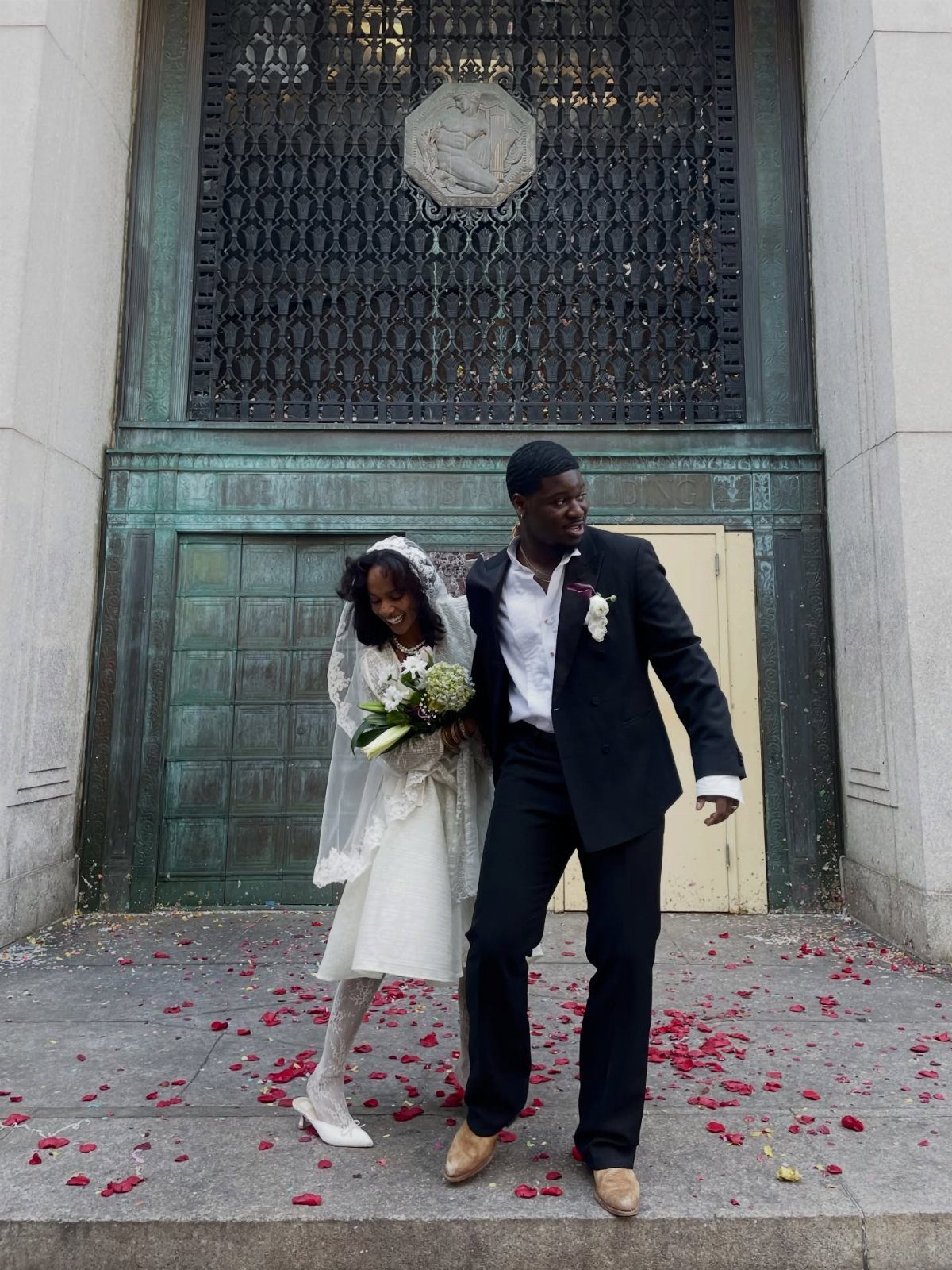 A bride in a vintage white wedding dress and veil walks with her groom outdoors.