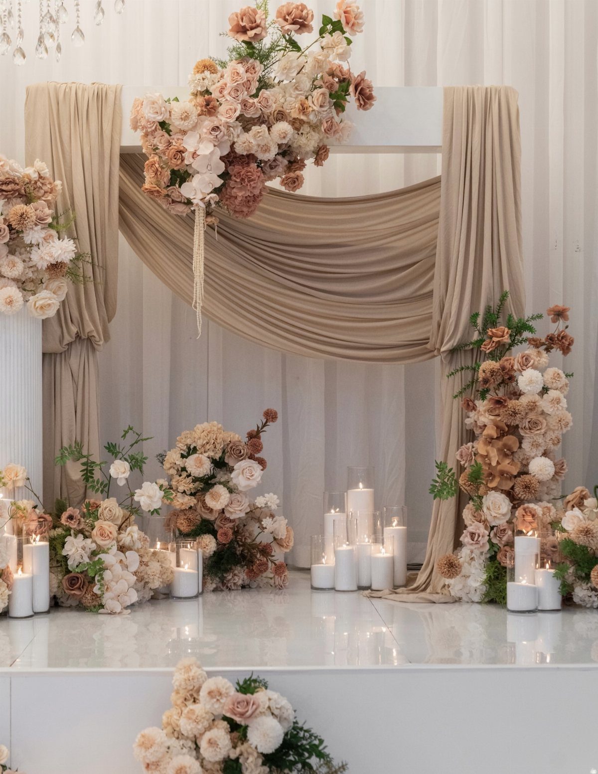 9 Earth Tone Wedding Colors That Feel Grounded, Not Dull 4 Wedding altar with beige draped fabric, peach floral arrangements, and white pillar candles on a white floor.
