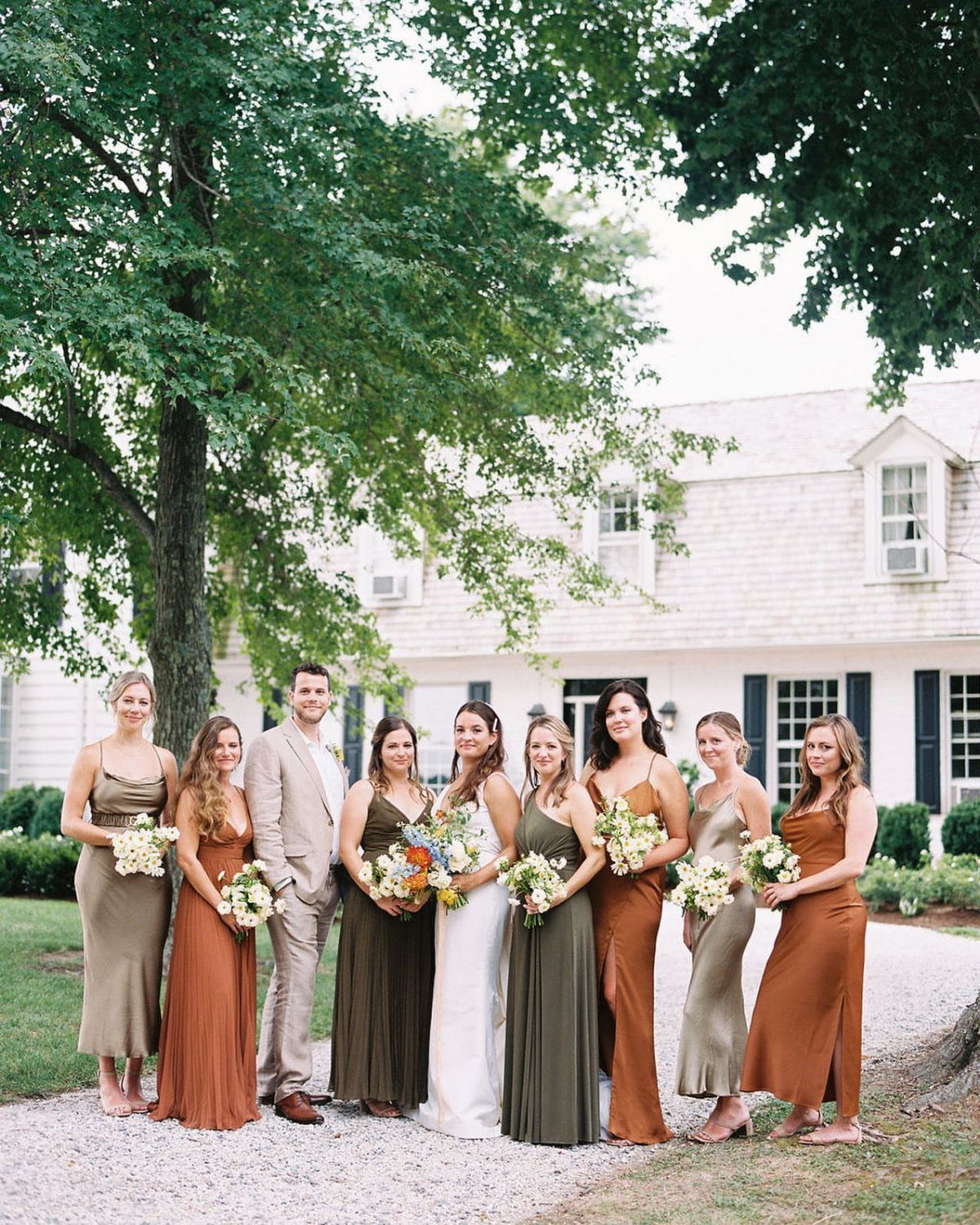 9 Earth Tone Wedding Colors That Feel Grounded, Not Dull 3 Bridesmaids in olive and terracotta dresses stand with a groom in front of a white house.