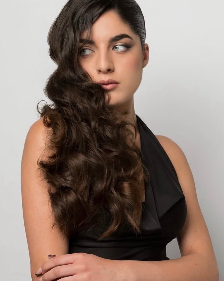 Woman with long dark hair styled in voluminous side-swept waves against a grey background.