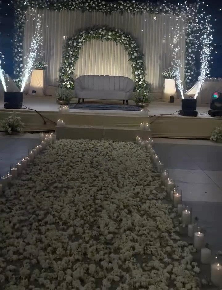 White flower petal aisle lined with small glowing candles leading to a floral archway.
