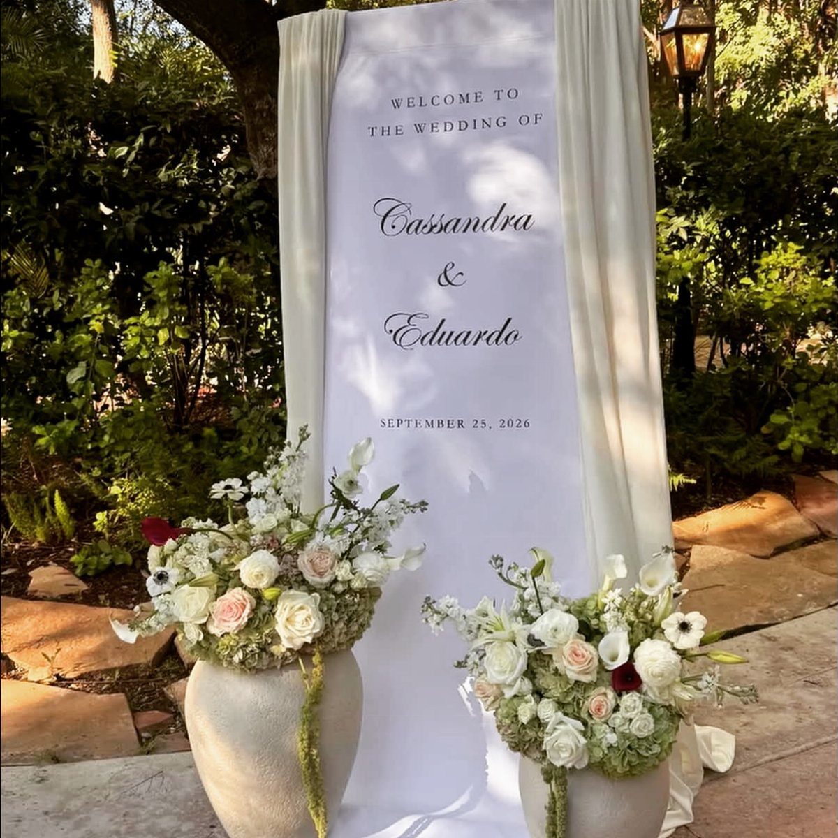 A white fabric wedding welcome sign flanked by cream drapes and floral arrangements.