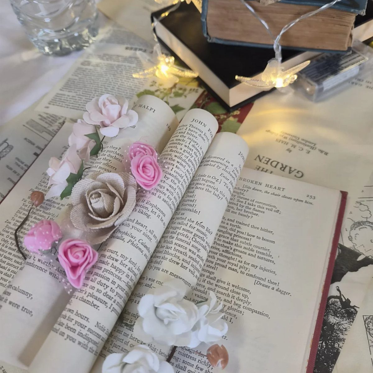 Paper scrolls decorated with fabric roses resting on printed pages and old books with fairy lights.