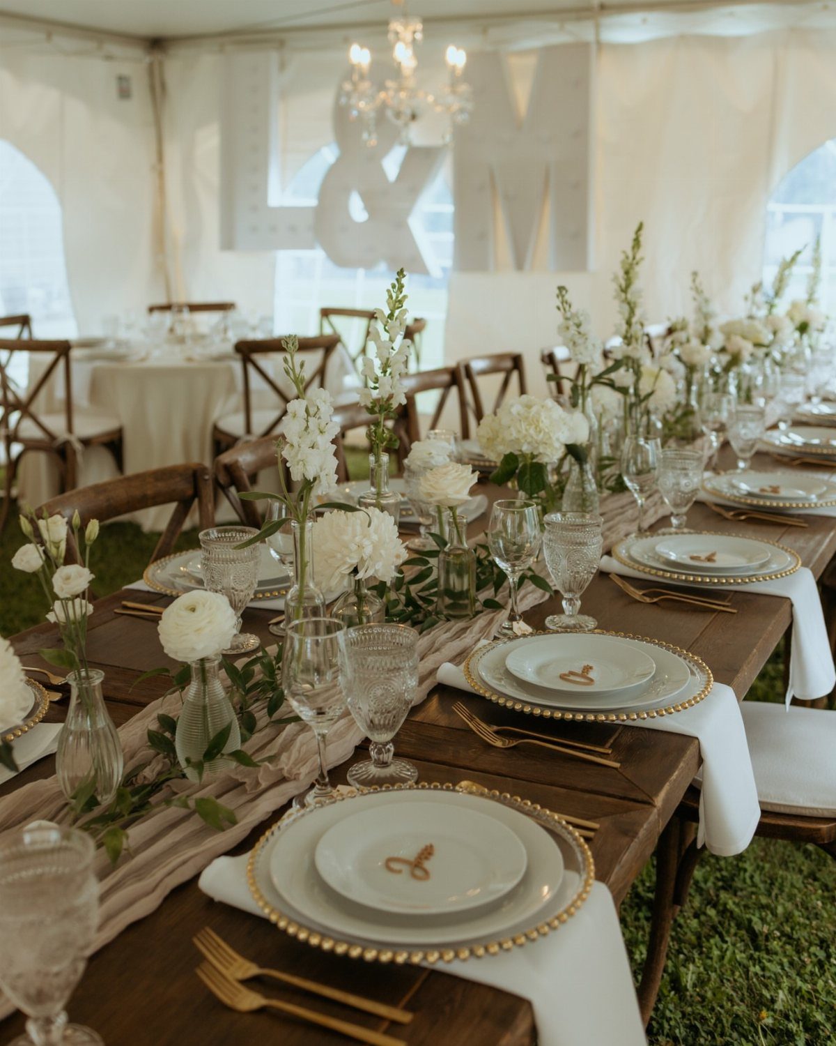Long wooden wedding table set with white plates, gold cutlery, and white floral centerpieces.