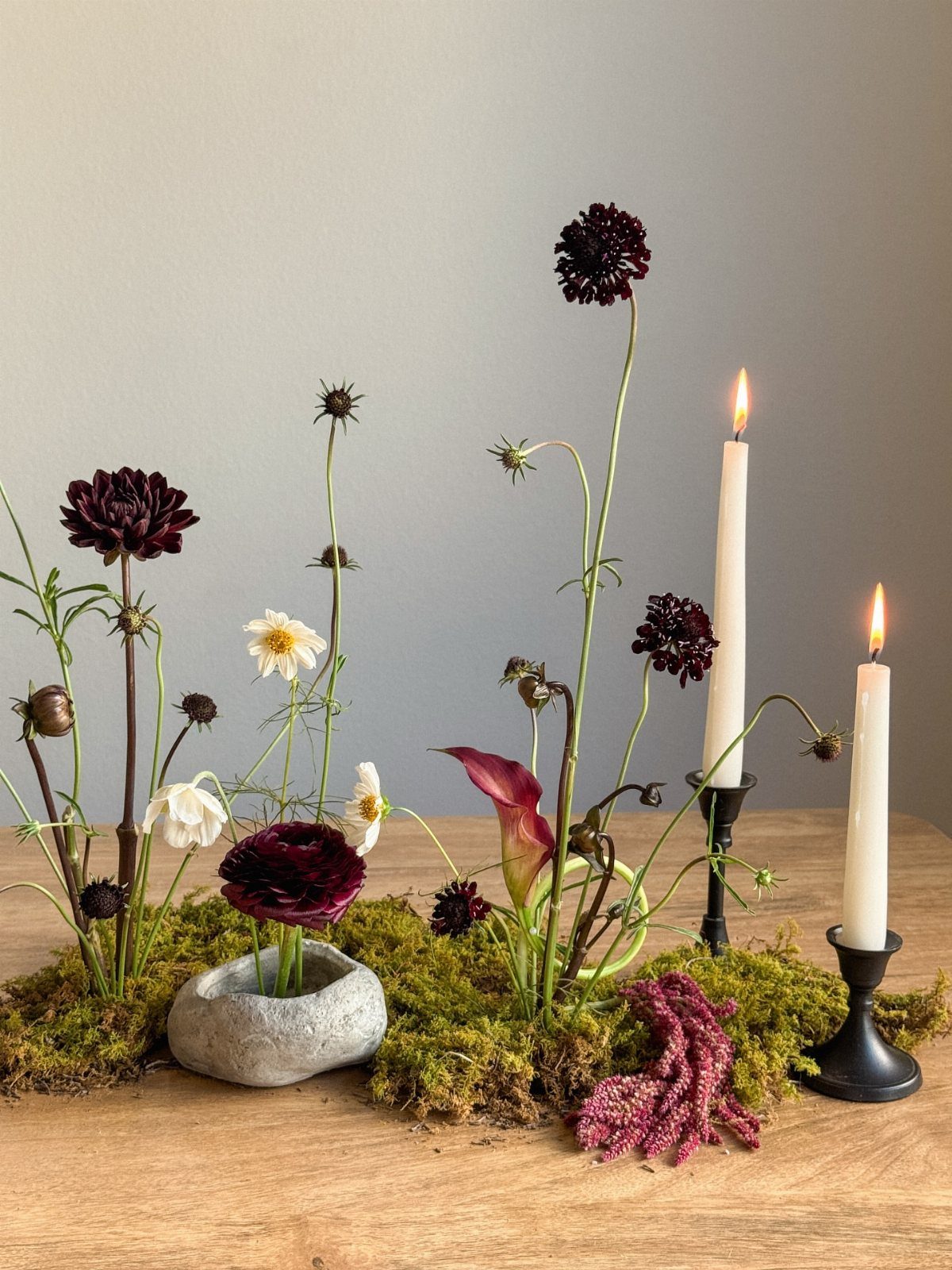 Moss table centerpiece with dark burgundy flowers, white blossoms, and lit taper candles on wood.