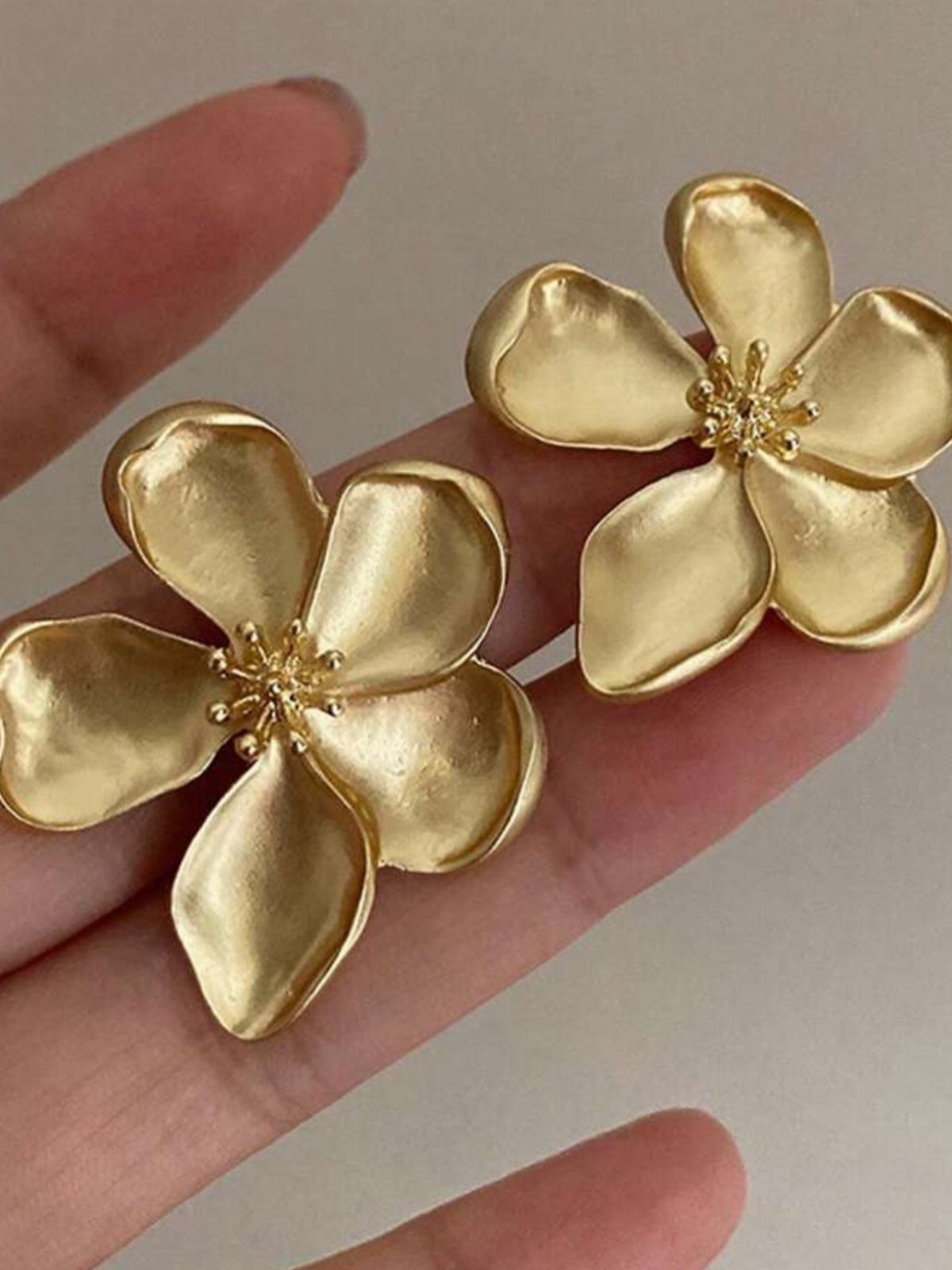 Two gold floral stud earrings held between a person's fingers against a neutral background.