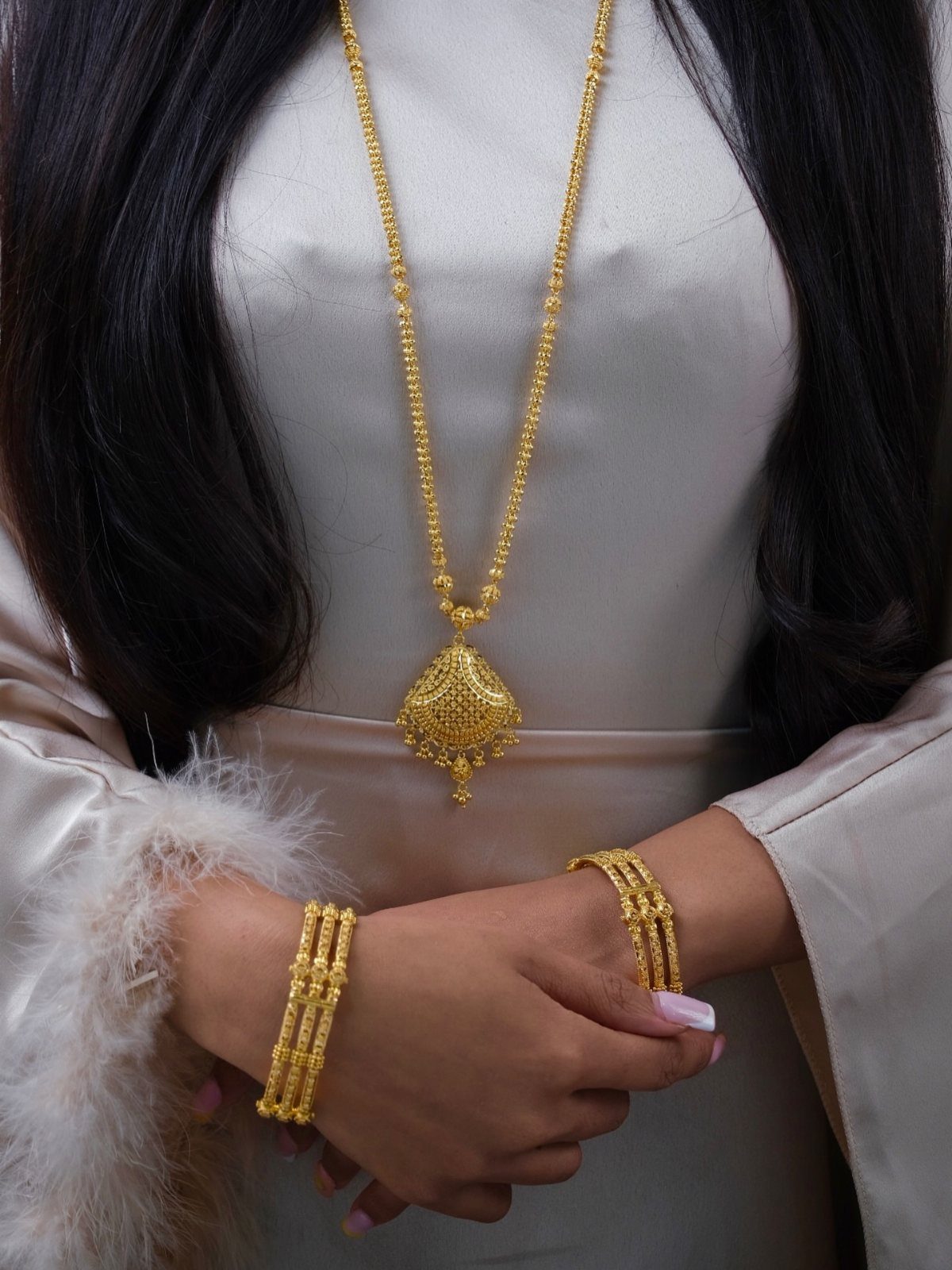 A person wearing a long gold chain necklace with a large ornate pendant and matching gold bangles.