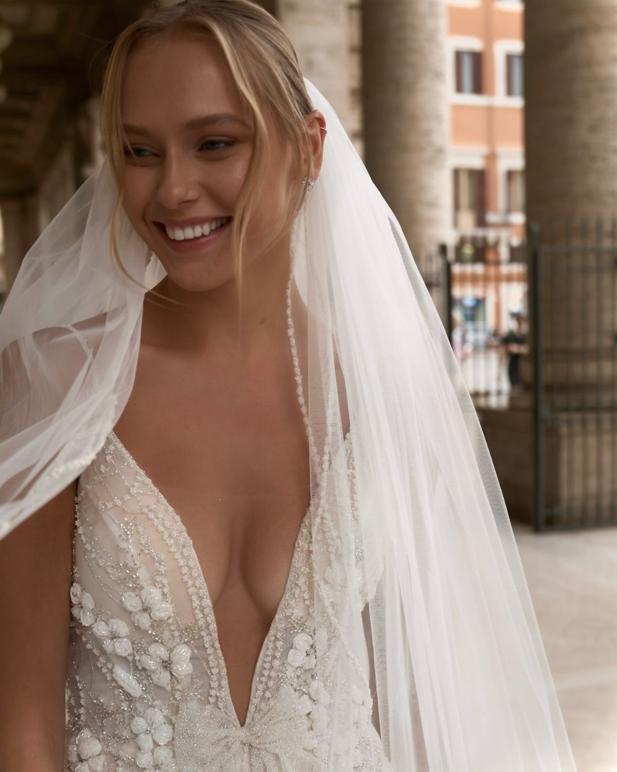 Smiling blonde woman wearing a white lace wedding dress with a deep plunging neckline and veil.