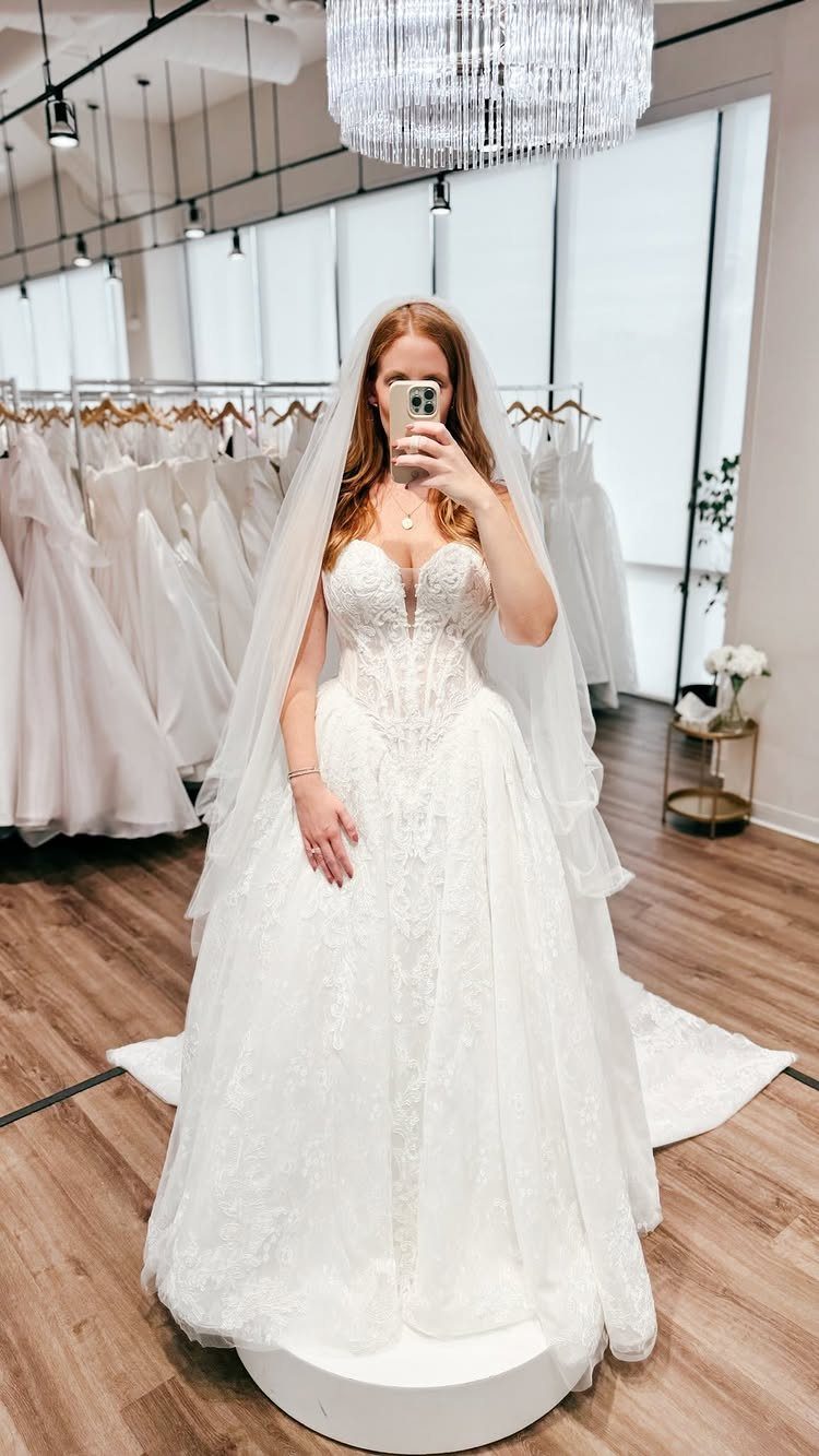 A woman wearing a white lace wedding dress and veil takes a mirror selfie in a bridal shop.