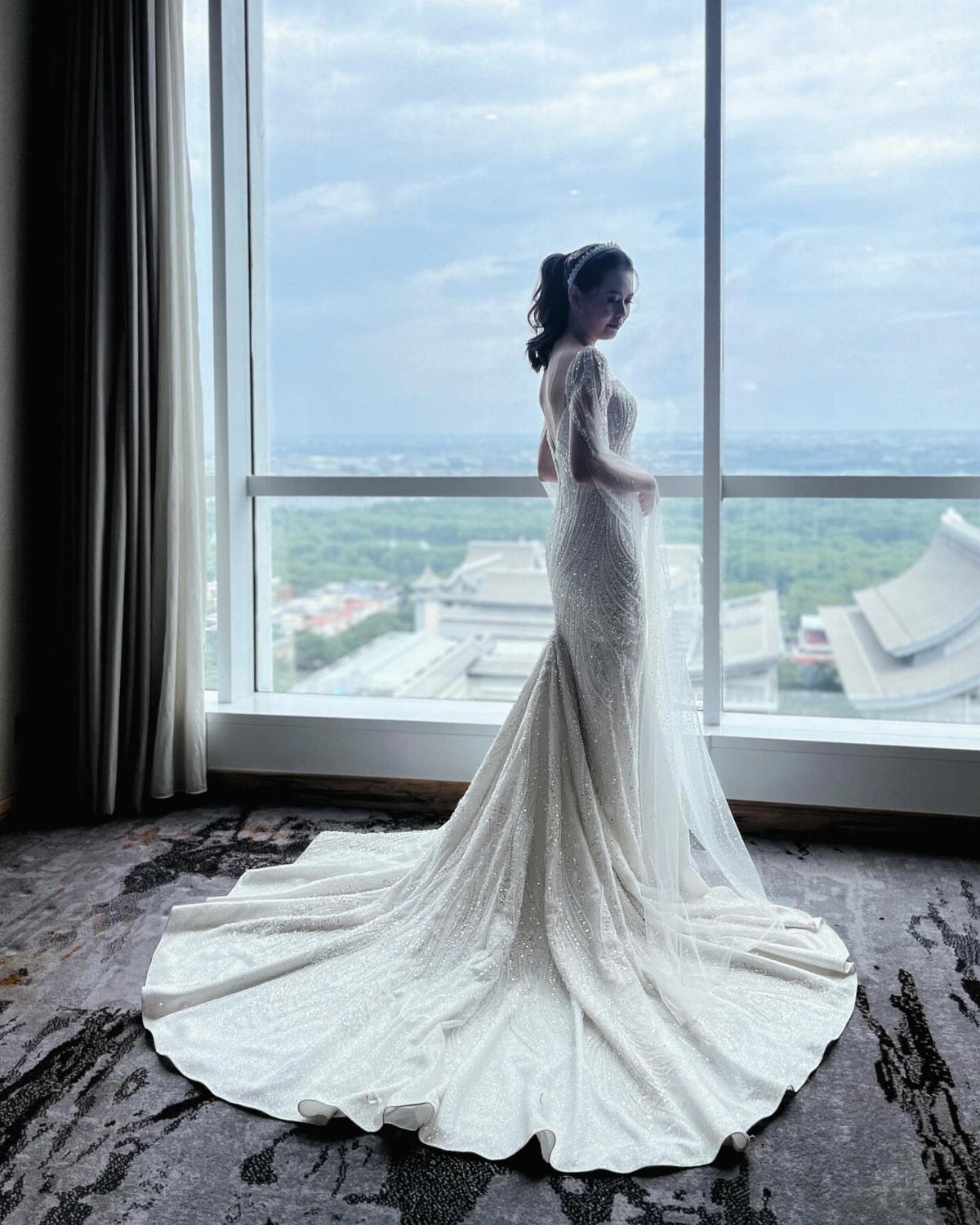 A woman wearing a sparkling white mermaid wedding gown standing by a large window.