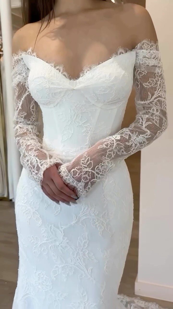 A woman wearing a white lace off-the-shoulder wedding gown with long sheer lace sleeves.