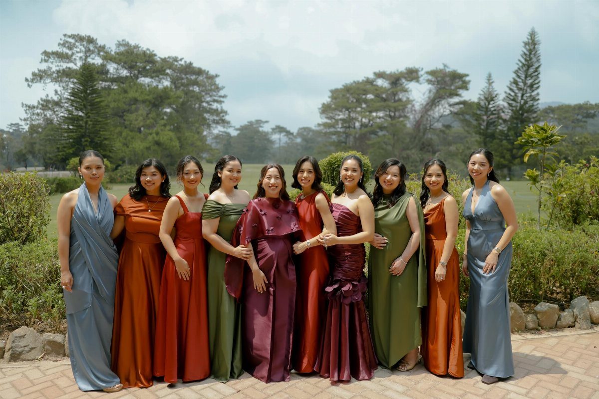 Ten women standing in a row wearing long jewel-toned evening gowns in an outdoor setting.